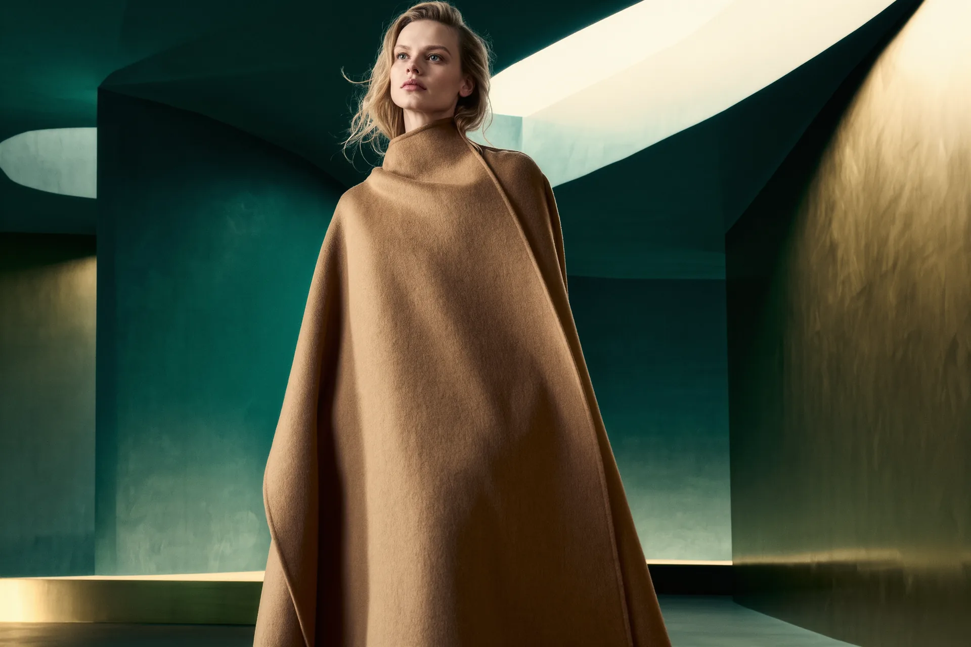 Un long manteau en cachemire camel posé sur une épaule, dans une lumière d'atelier tamisée.